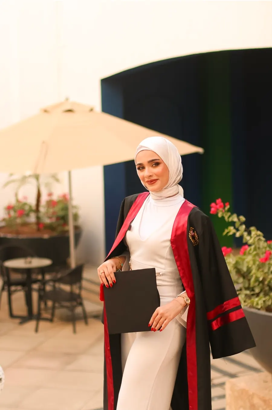 خرّيجة بروب التخرّج تحمل القبّعة بيديها في مساحة خارجية بمظلّة ونباتات